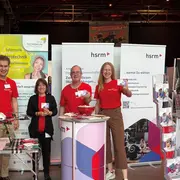 Auf dem Bild sind vier Personen in roten T-Shirts am Stand der Hochschule für Technik und Wirtschaft. Sie lächeln und halten Informationsmaterial in der Hand. Im Hintergrund sind Banner mit Informationen zur Hochschule zu sehen. Der Stand ist beleuchtet und freundlich gestaltet.