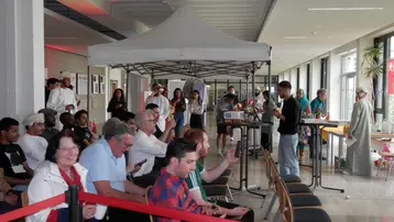 Eine Gruppe von Menschen sitzt in einem modernen Raum mit Tischen und Stühlen. Einige schauen auf ihre Handys, während andere miteinander sprechen. Im Hintergrund steht ein Zelt, und mehrere Personen scheinen an einem Event oder einer Veranstaltung teilzunehmen.