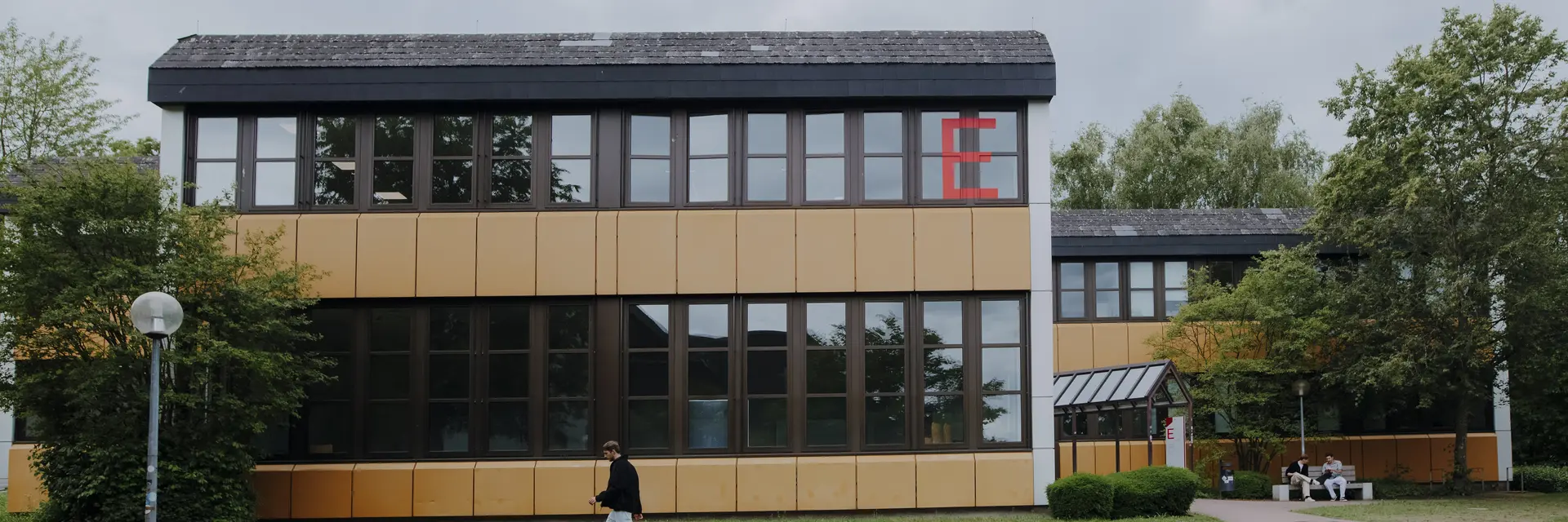 Ein modernes, zweigeschossiges Gebäude mit großen Fenstern und einer gelben Fassade. Vor dem Gebäude steht eine Person, die auf dem Gehweg entlanggeht. Auf der linken Seite sind Bäume und ein Lichtmast zu sehen. Am Gebäude ist ein großes rotes "E" zu sehen.