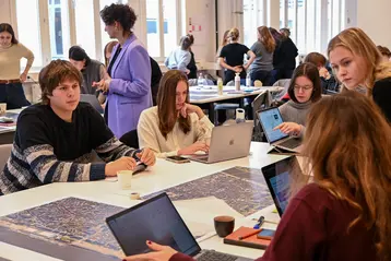 The image shows a group of international students at the University of Antwerp working at tables. Some are using laptops, while others are discussing. There is a large map on one table. The environment appears creative and collaborative, with several people in the background also working on projects. This is the HERILAND workshop.