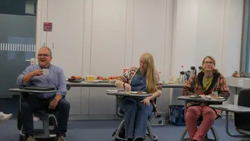 Drei Personen sitzen an Tischen in einem hellen Raum. Ein Mann lacht und hält eine Tasse. Eine Frau mit langen, blonden Haaren isst und eine weitere Frau mit brünetten Haaren schaut nachdenklich. Im Hintergrund sind Snacks und Getränke auf einem Tisch zu sehen.