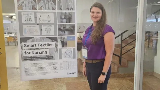 Eine Frau mit langen, braunen Haaren steht neben einem Plakat mit dem Titel „Smart Textiles for Nursing“. Das Plakat enthält Bilder und Informationen über innovative Textilien im Pflegebereich. Die Frau trägt ein lila Oberteil und hat eine Namensschilderung um ihren Hals.