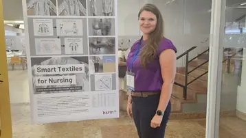 Eine Frau mit langen, braunen Haaren steht neben einem Plakat mit dem Titel „Smart Textiles for Nursing“. Das Plakat enthält Bilder und Informationen über innovative Textilien im Pflegebereich. Die Frau trägt ein lila Oberteil und hat eine Namensschilderung um ihren Hals.