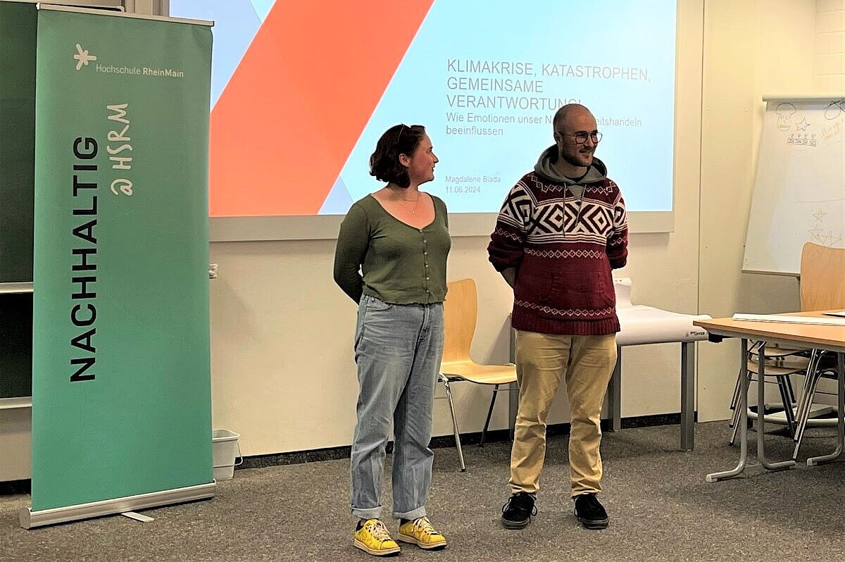 Zwei studentische Mitarbeitende des Nachhaltigkeitsbüros stehen vor den Anwesenden, neben ihnen steht auf einem Banner "Nachhaltig @ HSRM"