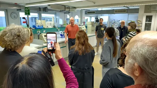 Eine Gruppe von Menschen steht in einem Labor. Ein Mann in einem roten Shirt spricht zu ihnen, während einige Zuhörer ihn mit ihren Handys aufnehmen. Die Atmosphäre ist interaktiv und informativ, mit wissenschaftlichen Geräten im Hintergrund.
