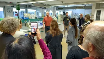 Eine Gruppe von Menschen steht in einem Labor. Ein Mann in einem roten Shirt spricht zu ihnen, während einige Zuhörer ihn mit ihren Handys aufnehmen. Die Atmosphäre ist interaktiv und informativ, mit wissenschaftlichen Geräten im Hintergrund.