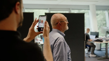 Eine Person filmt mit einem Smartphone eine andere Person, die im Vordergrund steht. Der Aufgenommene hat graue Haare und trägt ein blaues Hemd. Im Hintergrund sind Büroarbeitsplätze sichtbar.