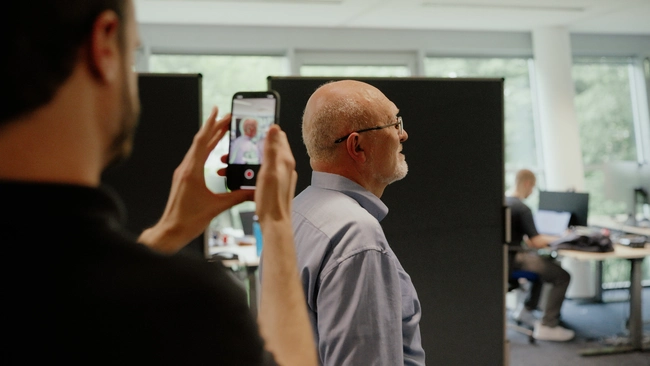 Eine Person filmt mit einem Smartphone eine andere Person, die im Vordergrund steht. Der Aufgenommene hat graue Haare und trägt ein blaues Hemd. Im Hintergrund sind Büroarbeitsplätze sichtbar.