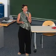 Eine Frau steht an einem Rednerpult und spricht zu einem Publikum. Sie trägt ein graues Top und hat die Haare kurz. Im Hintergrund sind ein Projektor und Tische sichtbar. Die Zuhörer sitzen auf Stühlen und einige halten Smartphones hoch.