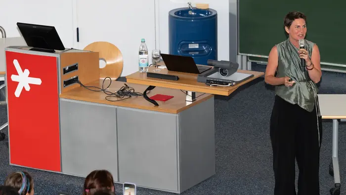 Eine Frau steht an einem Rednerpult und spricht zu einem Publikum. Sie trägt ein graues Top und hat die Haare kurz. Im Hintergrund sind ein Projektor und Tische sichtbar. Die Zuhörer sitzen auf Stühlen und einige halten Smartphones hoch.