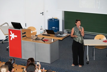 Eine Frau steht an einem Rednerpult und spricht zu einem Publikum. Sie trägt ein graues Top und hat die Haare kurz. Im Hintergrund sind ein Projektor und Tische sichtbar. Die Zuhörer sitzen auf Stühlen und einige halten Smartphones hoch.