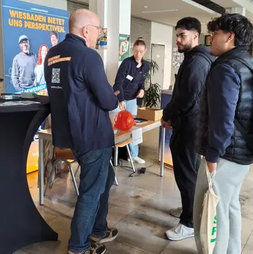 Eine Gruppe von drei Personen steht an einem Tisch. Ein Mann mit Brille spricht mit zwei jungen Männern, während eine Frau im Hintergrund beobachtet. Auf einem Banner im Hintergrund steht "Wiesbaden bietet uns Perspektiven". Der Raum hat eine helle, freundliche Atmosphäre.
