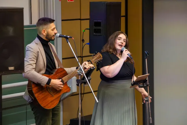 Ein Mann mit einer Gitarre und eine Frau mit Mikrofon stehen auf einer Bühne. Der Mann trägt einen grauen Anzug und die Frau ein schwarzes Oberteil mit einem langen Rock. Beide singen und scheinen eine musikalische Darbietung zu geben.