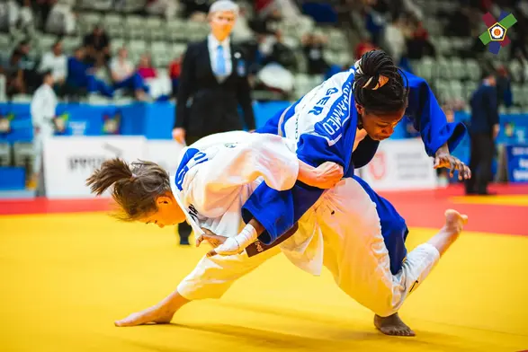 Zwei Judo-Kämpferinnen befinden sich in einem spannenden Wettkampf. Eine Kämpferin in blauer Kleidung führt einen Wurf aus, während die andere in weißer Kleidung nach unten sinkt. Der Hintergrund zeigt eine Sportarena mit Zuschauer:innen.