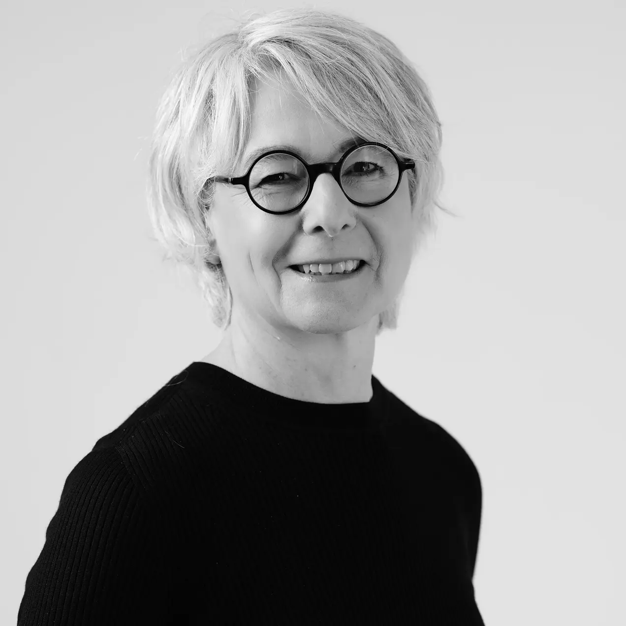 Eine lächelnde Frau mit kurzen, blonden Haaren und einer schwarzen, langen Armshirt steht vor einem hellen Hintergrund. Sie trägt eine Brille mit runden, schwarzen Rahmen und wirkt freundlich und selbstbewusst.