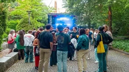 Eine Gruppe junger Menschen versammelt sich in einem Garten, umgeben von Bäumen. Im Hintergrund steht eine Bühne mit Lichtern und einem DJ. Die Atmosphäre wirkt entspannt und gesellig, während die Leute miteinander kommunizieren und die Musik genießen.