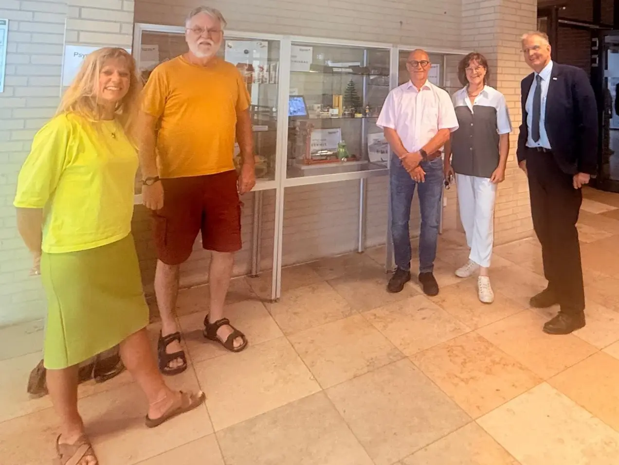 Fünf Personen stehen zusammen in einem Innenraum vor einer Vitrine. Die Gruppe besteht aus zwei Frauen und drei Männern, die in verschiedenen farbigen Outfits gekleidet sind. Die Vitrine im Hintergrund enthält verschiedene Ausstellungsstücke.