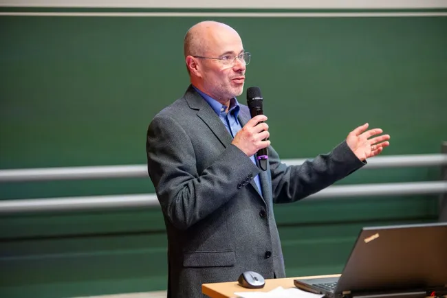 Ein Referent steht an einem Tisch und hält einen Vortrag. Er trägt einen grauen Anzug und spricht mit einem Mikrofon in der Hand. Auf dem Tisch befindet sich ein Laptop. Der Hintergrund ist grün und schlicht, was die Aufmerksamkeit auf den Sprecher lenkt.