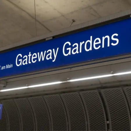 Schild mit der Aufschrift „Gateway Gardens“ und „Frankfurt am Main“. Es befindet sich an einem Bahnhof und zeigt eine Haltestelle an. Unter dem Hauptschild ist eine kleine Anzeigetafel mit „C 2“.