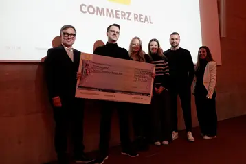 Eine Gruppe von fünf Personen steht lächelnd vor einem Bildschirm mit dem Logo "Commerz Real". Terence Reuschel hält einen großen Scheck in der Hand, der einen Betrag von 1.000 Euro zeigt. Er erhielt den Schwerpunktpreis im Bereich Digitalisierung im Bauprojekt- und Gebäudemanagement.