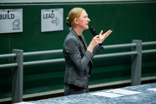 Eine Frau in einem grauen Anzug steht an einem Tisch und spricht mit Mikrofon. Sie gestikuliert mit ihren Händen, während sie auf ein Publikum hinweist. Im Hintergrund sind Schilder mit den Worten "BUILD" und "LEAD" zu sehen.