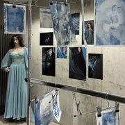 Eine Ausstellung mit Bildern in verschiedenen Blau- und Grautönen, die emotionale Porträts und Mode zeigen. Eine Schaufensterpuppe in einem langen, hellblauen Kleid steht im Vordergrund, während gedruckte Bilder an einer Wand befestigt sind. Die Atmosphäre ist künstlerisch und kreativ.