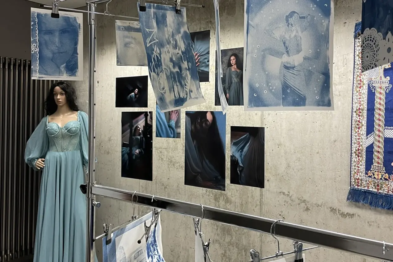 Eine Ausstellung mit Bildern in verschiedenen Blau- und Grautönen, die emotionale Porträts und Mode zeigen. Eine Schaufensterpuppe in einem langen, hellblauen Kleid steht im Vordergrund, während gedruckte Bilder an einer Wand befestigt sind. Die Atmosphäre ist künstlerisch und kreativ.