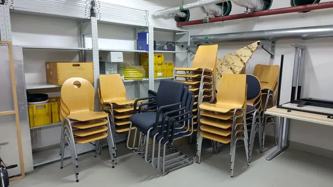 In einem Raum stehen mehrere gestapelte Holzstühle. In der Mitte befindet sich ein blauer Stuhl, der anders angeordnet ist. Regale an der Wand enthalten verschiedene Kisten und Materialien. Der Boden ist leicht grau und die Umgebung wirkt aufgeräumt.