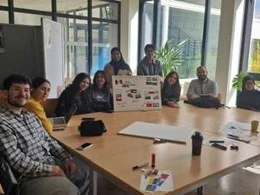 Eine Gruppe von zehn Personen sitzt an einem Tisch in einem hellen Raum. Sie arbeiten an einem Projekt, das auf einem großen Plakat präsentiert wird. Auf dem Tisch liegen Materialien wie Marker und Notizen, während einige Teilnehmer in entspannter Atmosphäre diskutieren.