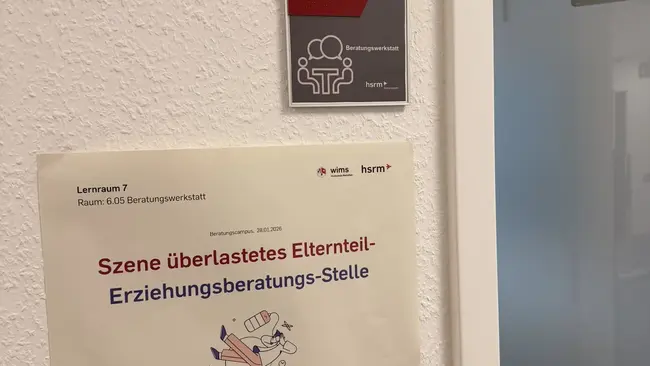 Ein Schild an einer Wand zeigt den Raum B 6.05, gekennzeichnet mit einem Piktogramm für Menschen. Darunter befindet sich ein weiteres Schild, auf dem steht: "Szene überlastetes Elternteil - Erziehungsberatungsstelle". Die Umgebung hat einen neutralen, modernen Charakter.