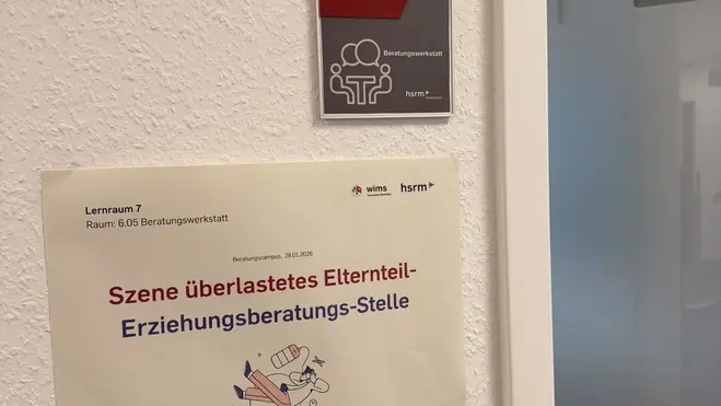 Ein Schild an einer Wand zeigt den Raum B 6.05, gekennzeichnet mit einem Piktogramm für Menschen. Darunter befindet sich ein weiteres Schild, auf dem steht: "Szene überlastetes Elternteil - Erziehungsberatungsstelle". Die Umgebung hat einen neutralen, modernen Charakter.