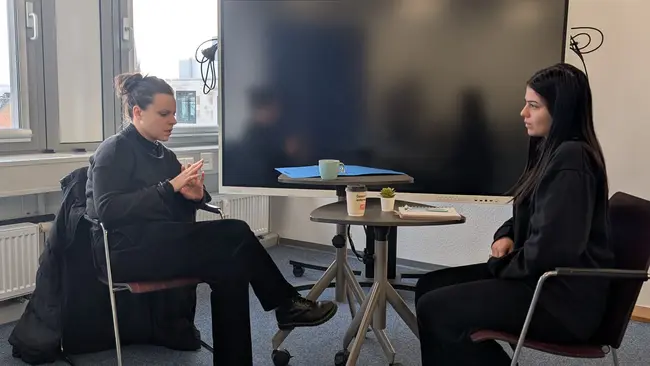 Zwei Personen sitzen an einem Tisch in einem Büro. Eine Person hat ein Smartphone in der Hand und schaut darauf, während die andere Person sie ansieht. Vor ihnen steht ein Tisch mit Getränken. Im Hintergrund ist ein großer Bildschirm zu sehen.