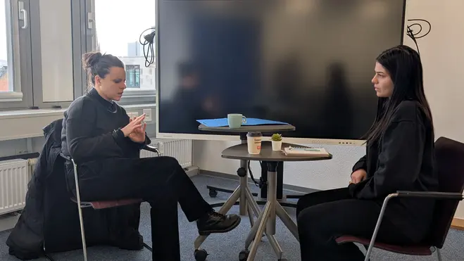 Zwei Personen sitzen an einem Tisch in einem Büro. Eine Person hat ein Smartphone in der Hand und schaut darauf, während die andere Person sie ansieht. Vor ihnen steht ein Tisch mit Getränken. Im Hintergrund ist ein großer Bildschirm zu sehen.