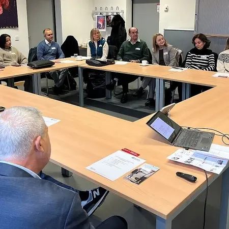 Eine Gruppe von Menschen sitzt um einen großen Konferenztisch. In der Mitte des Tisches steht ein Laptop, und auf dem Tisch liegen Unterlagen. Die Teilnehmer konzentrieren sich aufeinander und scheinen an einem Meeting oder einer Diskussion teilzunehmen.