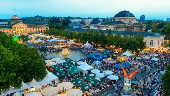 Ein lebhaftes Fest mit vielen Menschen, Verkaufsständen und Zelten. Im Hintergrund sind historische Gebäude zu sehen, umgeben von Bäumen. Die Atmosphäre wirkt festlich und einladend, mit stimmungsvollem Licht und einer großen Menschenmenge, die den Platz belebt.