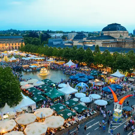 Ein lebhaftes Fest mit vielen Menschen, Verkaufsständen und Zelten. Im Hintergrund sind historische Gebäude zu sehen, umgeben von Bäumen. Die Atmosphäre wirkt festlich und einladend, mit stimmungsvollem Licht und einer großen Menschenmenge, die den Platz belebt.