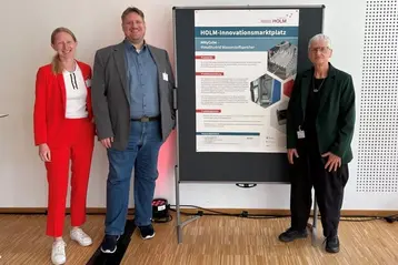 Drei Personen stehen neben einem Präsentationsposter mit dem Titel „HOLM-Innovationsmarktplatz“. Die Person links trägt einen roten Anzug, die in der Mitte einen grauen Anzug, und die rechts eine dunkle Jacke. Das Poster enthält Informationen zur Forschungs- und Innovationsarbeit.