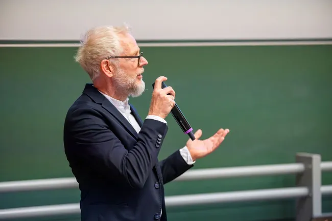 Ein älterer Mann mit grauem Haar und Brille steht vor einer grünen Tafel. Er hält ein Mikrofon in der Hand und spricht mit engagierter Gestik, während er seine andere Hand hebt. Sein Outfit ist formell, bestehend aus einem dunklen Anzug und einem hellen Hemd.