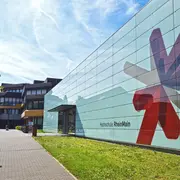 Eine moderne Hochschulgebäude mit einer großen, bunten Fassadengestaltung in Form von geometrischen Formen. Der Eingangsbereich ist gut sichtbar und führt über einen Gehweg, der von Grünflächen umgeben ist. Im Hintergrund sind weitere Gebäudeteile der Hochschule erkennbar.