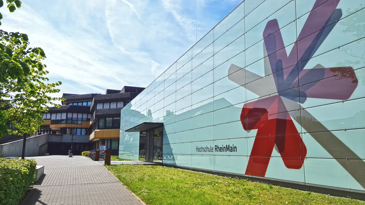 Eine moderne Hochschulgebäude mit einer großen, bunten Fassadengestaltung in Form von geometrischen Formen. Der Eingangsbereich ist gut sichtbar und führt über einen Gehweg, der von Grünflächen umgeben ist. Im Hintergrund sind weitere Gebäudeteile der Hochschule erkennbar.