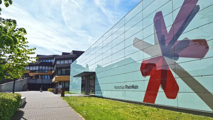 Eine moderne Hochschulgebäude mit einer großen, bunten Fassadengestaltung in Form von geometrischen Formen. Der Eingangsbereich ist gut sichtbar und führt über einen Gehweg, der von Grünflächen umgeben ist. Im Hintergrund sind weitere Gebäudeteile der Hochschule erkennbar.