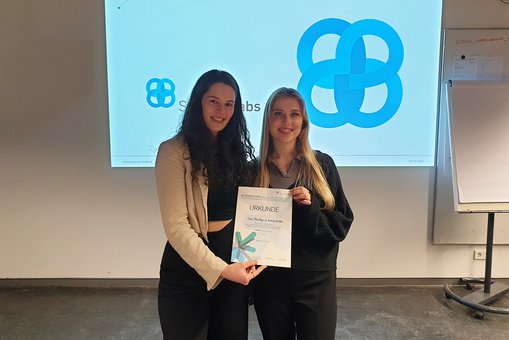 Die Kommunikationsdesign-Studentinnen Sara Boettger und Emily Endler stehen mit der Urkunde für den 3. Platz vor ihrem Logoentwurf