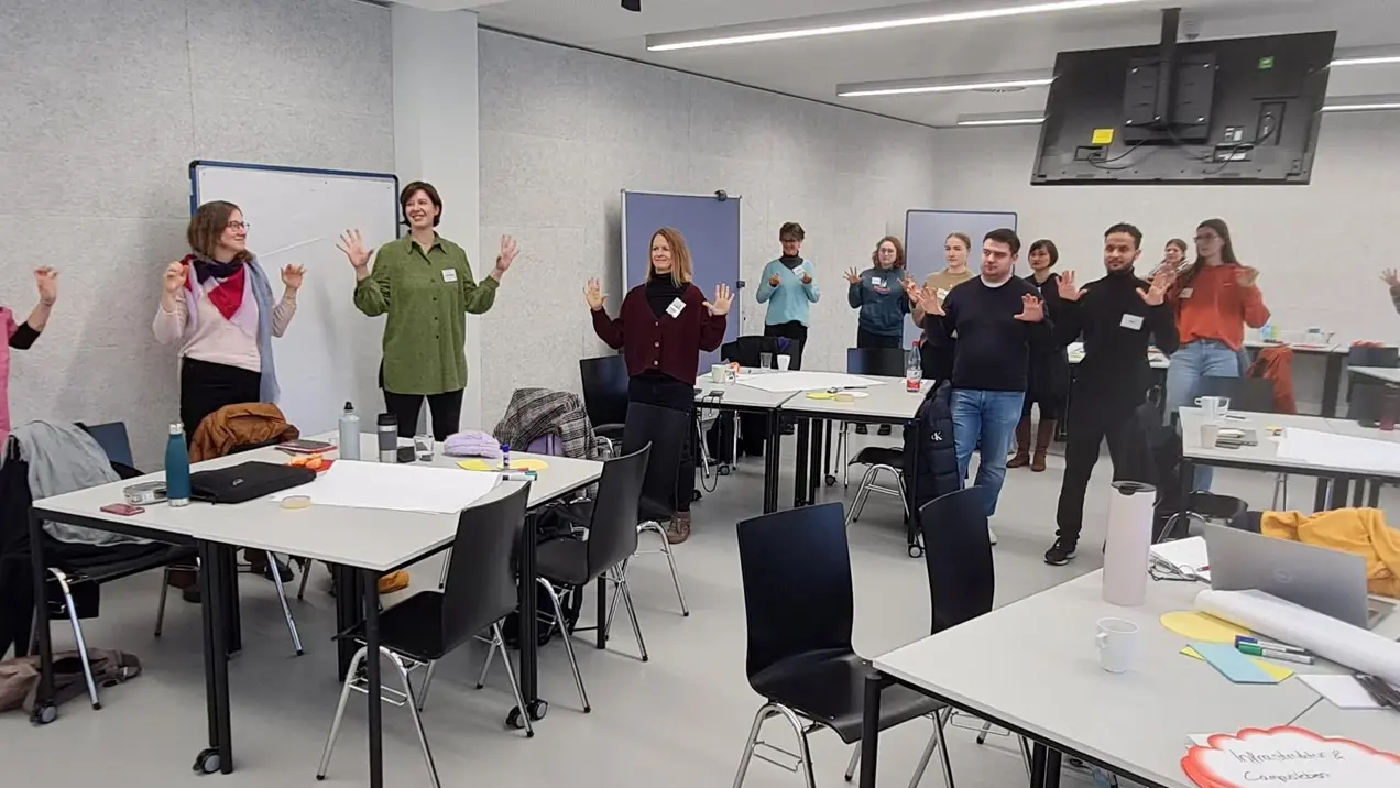 Eine Gruppe von Menschen steht in einem Seminarraum und hebt die Hände, als würde sie aktiv teilnehmen. Es sind mehrere Tische mit Stühlen im Hintergrund zu sehen. Alle Teilnehmer tragen Namensschilder. Der Raum wirkt hell und modern.