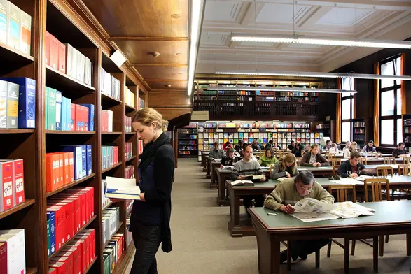 Studierende blättert in einem Buch vor einem Regal im Lesesaal in der Bibliothek