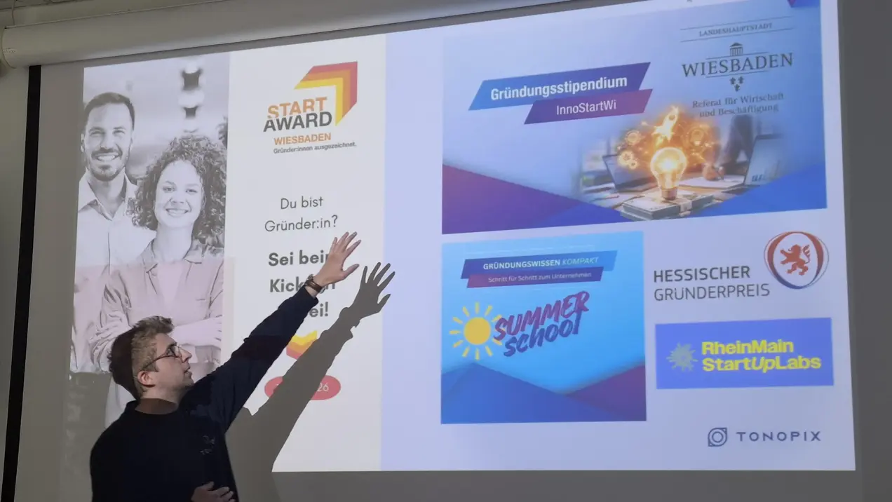 Ein Präsentator zeigt auf eine Folie, die Informationen über verschiedene Gründerstipendien und Wettbewerbe enthält. Der Text ermutigt Gründerinnen mit dem Hinweis: „Du bist Gründerin? Sei bold! Klicke hier!“ Die Folie enthält logos von Veranstaltungen in Wiesbaden und anderen Initiativen.