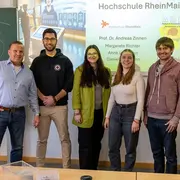 Eine Gruppe von fünf Personen steht lächelnd vor einer Präsentation im Hintergrund. Links steht ein Mann in Hemd, daneben ein Mann in einem schwarzen Hoodie. Die drei Frauen in der Mitte und rechts tragen lässige Kleidung. Im Hintergrund ist ein Logo und der Name "Hochschule RheinMain" zu sehen.