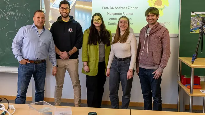 Eine Gruppe von fünf Personen steht lächelnd vor einer Präsentation im Hintergrund. Links steht ein Mann in Hemd, daneben ein Mann in einem schwarzen Hoodie. Die drei Frauen in der Mitte und rechts tragen lässige Kleidung. Im Hintergrund ist ein Logo und der Name "Hochschule RheinMain" zu sehen.