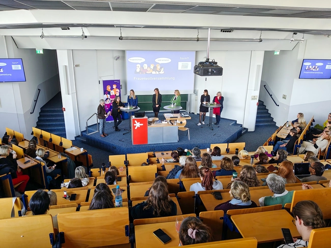 Eine Gruppe von fünf Personen steht auf einer Bühne vor einem Publikum in einem Konferenzraum. Die Präsentation zeigt diverse Themen. Zuschauer sitzen in Reihen, konzentriert auf die Diskussion. Ein Flipchart und mehrere Bildschirme sind ebenfalls sichtbar, um das Thema zu unterstützen.