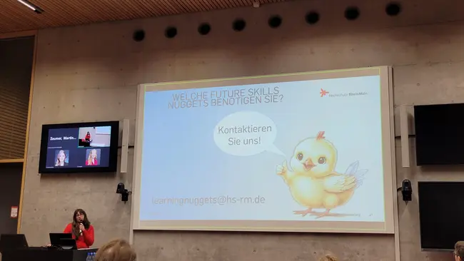 Eine Präsentation in einem modernen Raum, in der eine Person spricht. Auf der Leinwand steht: "Welche Future Skills Nuggets benötigen Sie? Kontaktieren Sie uns!" Neben dem Text ist ein niedliches, cartoonhaftes Küken abgebildet.