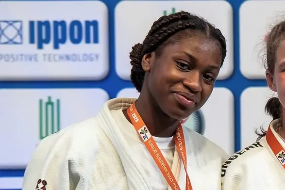 Eine junge Judoka trägt eine Medaille um den Hals und lächelt. Sie hat geflochtene Haare und steht neben einer anderen Sportlerin. Im Hintergrund sind Sponsorenlogos sichtbar. Die Atmosphäre wirkt feierlich und triumphierend.